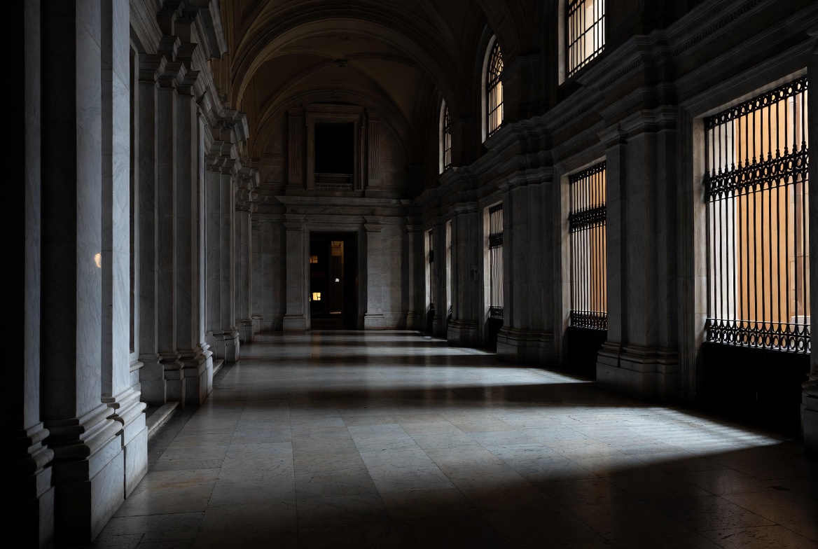 Empty marble corridor of a grand European institution — amber light through iron-barred windows, long shadows across stone floors. The weight of the monetary system made visible.
